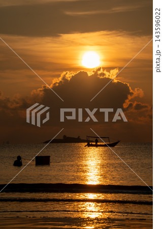 Captivating golden sunset over a tropical sea, featuring the sun reflecting on the calm water. Silhouettes of a boat, a person wading, and a distant island create a serene scene. 143596022