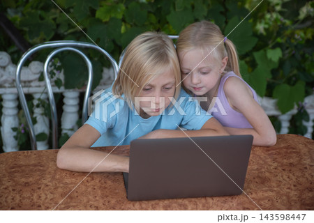 Young boy and girl sharing a laptop, focusing on the screen while engaging in a shared educational or entertainment activity on a patio with a leafy green background 143598447