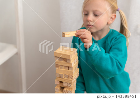 Child carefully placing a wooden block on top of a towering structure, developing concentration, fine motor skills, and problem solving abilities during playtime 143598451