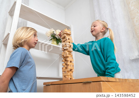 Young children playing a stacking blocks game at home, carefully removing a wooden piece from the high tower, highlighting concepts of balance, challenge, and focus during playtime 143598452