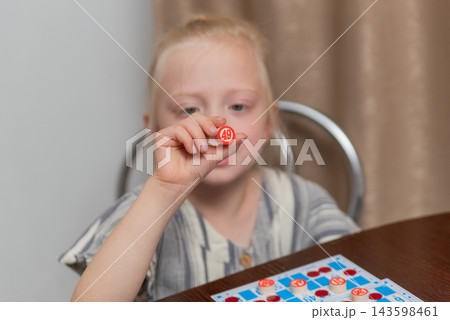 Child concentrating on the game, holding a red number token with 49 on it, participating in a fun family activity with bingo cards visible on the table 143598461