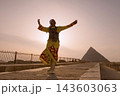 Traditional dancer in vibrant yellow embroidered robe spinning arms outstretched at iconic Giza Pyramids against glowing hazy sunset sky ideal for 143603063