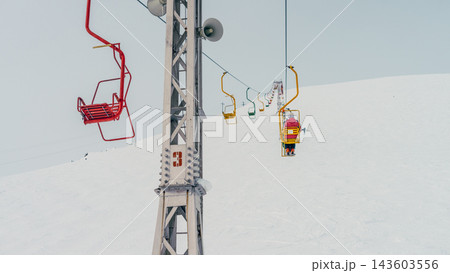 Skier on yellow chairlift ascending snowy slope past red empty seat and metal support tower Skier on yellow chairlift ascending snowy slope past red empty seat and metal support tower 143603556