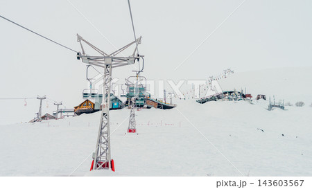Chairlift towers and empty chairs ascending snowy slope toward ski station buildings on pale winter day 143603567