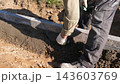 Construction workers spread wet concrete beside freshly set curb edging on sunlit soil, using gloves and hand tools to shape foundation edge at outdoor building site with close ground level view. 143603769