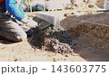 Construction worker kneels beside foundation trench, spreading and smoothing fresh concrete with trowel to form straight edge for residential house project in bright daylight outdoors. 143603775