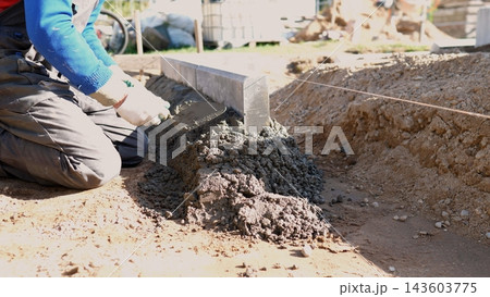 Construction worker kneels beside foundation trench, spreading and smoothing fresh concrete with trowel to form straight edge for residential house project in bright daylight outdoors. Construction worker kneels beside foundation trench, spreading and smoothing fresh concrete with trowel to form straight edge for residential house project in bright daylight outdoors. 143603775