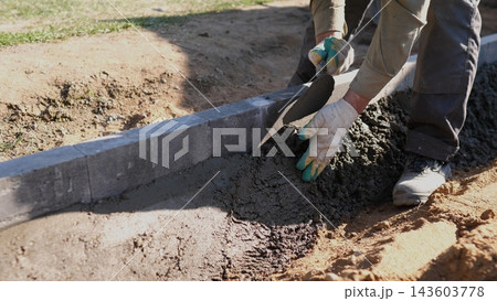 Worker spreads wet concrete along curb edge with trowel during outdoor construction, shaping fresh mortar beside dirt ground in bright daylight on residential site with close view. Worker spreads wet concrete along curb edge with trowel during outdoor construction, shaping fresh mortar beside dirt ground in bright daylight on residential site with close view. 143603778