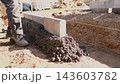 Worker leveling fresh concrete beside straight concrete edge in groundwork for house construction, with soil trench, guide string, boots, sunlight, and outdoor building site details. 143603782