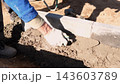 Construction worker wearing gloves levels wet mortar between concrete blocks in outdoor foundation trench, using hand trowel on dirt ground under bright sunlight and strong shadows. 143603789
