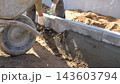 Close view of hands shaping wet concrete along curb edge at residential building site, with wheelbarrow, soil mound, and bright daylight highlighting masonry progress and careful finishing work. 143603794