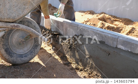 Close view of hands shaping wet concrete along curb edge at residential building site, with wheelbarrow, soil mound, and bright daylight highlighting masonry progress and careful finishing work. Close view of hands shaping wet concrete along curb edge at residential building site, with wheelbarrow, soil mound, and bright daylight highlighting masonry progress and careful finishing work. 143603794