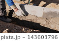 Worker smoothing fresh mortar beside concrete block foundation during house construction, with gloved hand, trowel, boots, trench, soil, and strong daylight at outdoor building site. 143603796