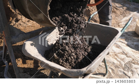Close view shows wet concrete spilling from mixer drum into wheelbarrow beside worker legs on dusty construction ground, with sunlight, tools, and foundation work materials visible. Close view shows wet concrete spilling from mixer drum into wheelbarrow beside worker legs on dusty construction ground, with sunlight, tools, and foundation work materials visible. 143603803