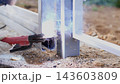 Close view of worker welding metal fencing outdoors, with bright arc light, flying sparks, smoke, protective gloves, and steel parts being joined on gravel ground in daylight 143603809