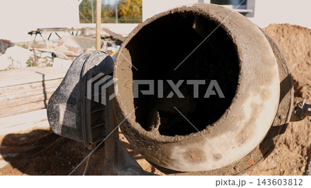 Mud covered concrete mixer drum at construction site, shown close up beside piles of soil and unfinished building materials in bright daylight, highlighting heavy work and building progress. 143603812