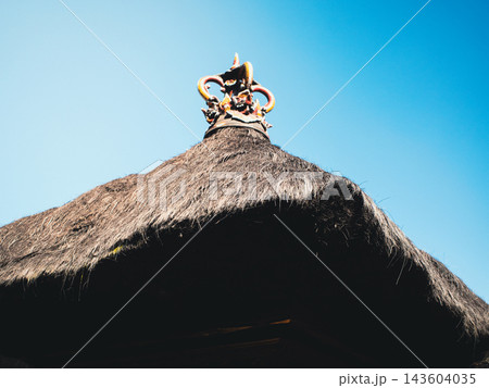 Balinese temple roof with layered thatch and ornate sacred ornament at the apex, set against a vivid blue sky — traditional Hindu architecture reflecting local craftsmanship and spirituality Balinese temple roof with layered thatch and ornate sacred ornament at the apex, set against a vivid blue sky — traditional Hindu architecture reflecting local craftsmanship and spirituality 143604035