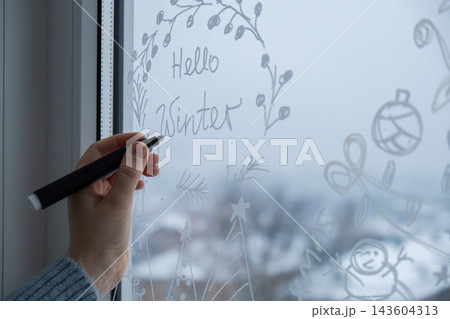 Sustainable responsible holiday decor. Hello winter text painted on window glass indoor at home. Christmas winter decoration. White pattern. Snow art tracery on season. Diy Sustainable responsible holiday decor. Hello winter text painted on window glass indoor at home. Christmas winter decoration. White pattern. Snow art tracery on season. Diy 143604313