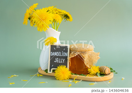 Transparent glass of jar DANDELION JELLY text with dandelion flowers in vase. Soft natural lighting highlights the amber color glossy texture and artisanal character of the preserve. Concept of 143604636
