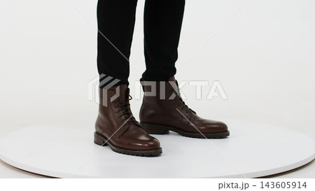 Person wearing brown leather lace-up boots and black trousers, showcasing male footwear fashion on a white background Person wearing brown leather lace-up boots and black trousers, showcasing male footwear fashion on a white background 143605914