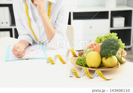 Nutritionist at desk with fruits, vegetables and measuring tape in clinic, closeup Nutritionist at desk with fruits, vegetables and measuring tape in clinic, closeup 143606110
