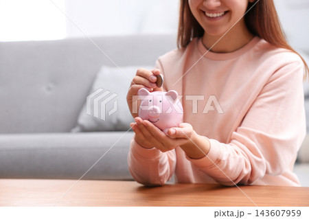 Woman putting coin into piggy bank at wooden table, closeup Woman putting coin into piggy bank at wooden table, closeup 143607959