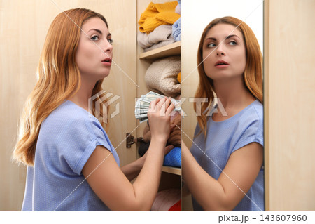 Worried woman hiding money between clothes in wardrobe indoors. Financial savings Worried woman hiding money between clothes in wardrobe indoors. Financial savings 143607960