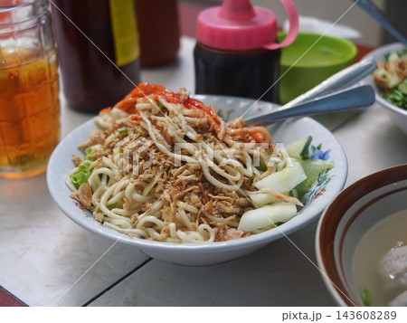 A bowl of Indonesian chicken noodles with red chili sauce on top alongside condiments and iced tea. 143608289
