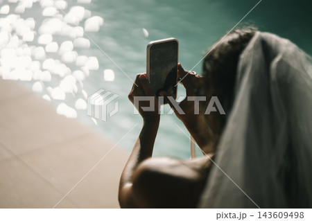Bride holding a smartphone beside a sunlit swimming pool 143609498