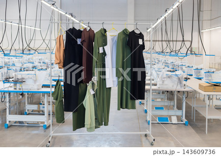 Clothes hanging on a rack in a clothing factory with sewing machines in the background 143609736
