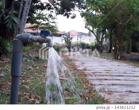 Water spraying from grey spigot. Close up view of splash in nature. Irrigation system in park setting. Walking trail background with leaves. Water spraying from grey spigot. Close up view of splash in nature. Irrigation system in park setting. Walking trail background with leaves. 143611212