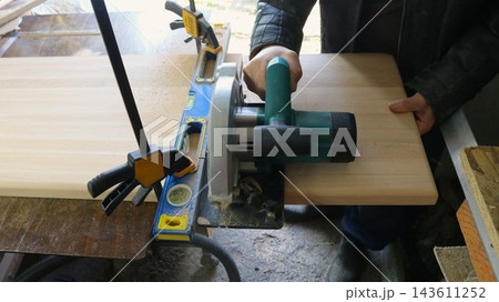 A carpenter advances a handheld circular saw across the wood grain, using a metal spirit level as a straight guide. Trimming the end of a solid wood tabletop with an electric circular saw A carpenter advances a handheld circular saw across the wood grain, using a metal spirit level as a straight guide. Trimming the end of a solid wood tabletop with an electric circular saw 143611252