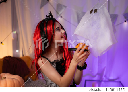 Cute Little Girl In A Halloween Witch Wig Plays With A Pumpkin, Drawing A Spooky Face And Showing It To A Ghost 143611325