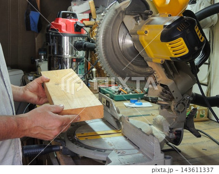 The craftsman holds a thick wooden block, positioning it against the bed of a miter saw to make a precise cut. A professional construction vacuum cleaner is visible in the background. The craftsman holds a thick wooden block, positioning it against the bed of a miter saw to make a precise cut. A professional construction vacuum cleaner is visible in the background. 143611337
