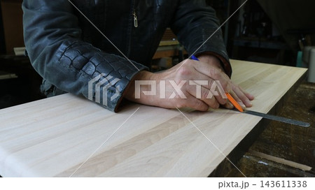 A worker in the workshop is marking the outlines of a part. A thin steel ruler lies on the surface of a light-colored panel, and a mark for the future cut is carefully drawn along it with a pen. 143611338