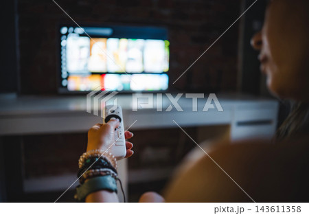 Young Woman In A Dark Room Watching A Smart Tv And Changing Channels With A Remote 143611358
