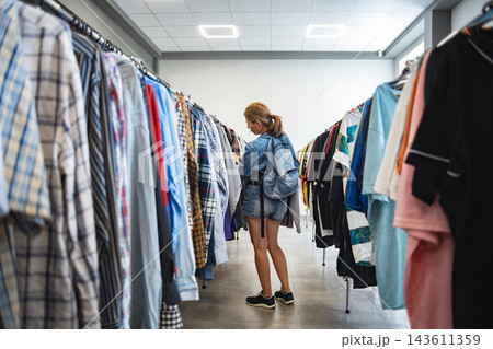 Redheaded young woman examines hanging garments, standing between two long rows of clothing racks in secondhand thrift shop 143611359