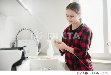 Girl washes a glass at the kitchen sink while wearing a red shirt. Washing glass with dish towel. 143611360