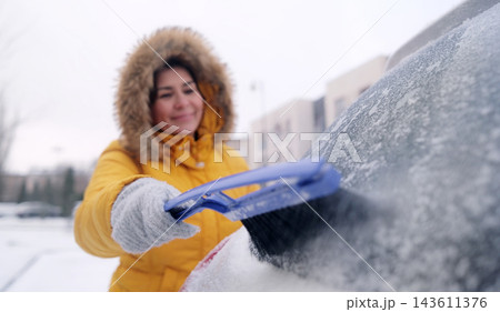 Woman in a yellow jacket removes snow using a brush on a cold day in a parking lot Woman in a yellow jacket removes snow using a brush on a cold day in a parking lot 143611376