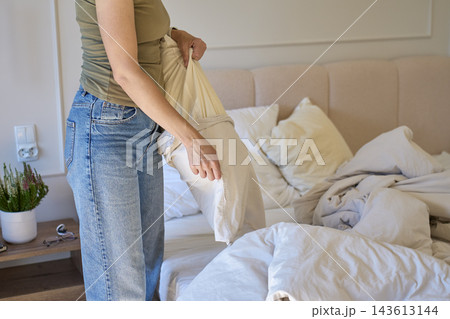 Woman pulling beige pillowcase off pillow beside unmade bed with white bedding 143613144