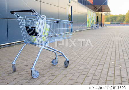 Metallic shopping cart filled with plastic grocery bag stands on paved parking lot outside supermarket against autumn backdrop Metallic shopping cart filled with plastic grocery bag stands on paved parking lot outside supermarket against autumn backdrop 143613293