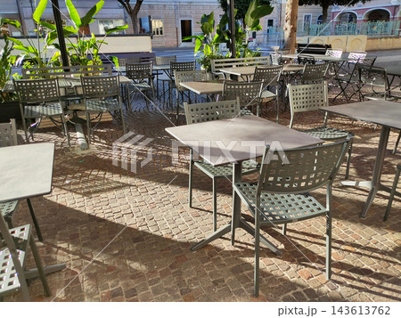 Outdoor cafe terrace with empty metal tables and chairs on a rustic cobblestone pavement, surrounded by tropical plants and historic European buildings in warm sunlight. Outdoor cafe terrace with empty metal tables and chairs on a rustic cobblestone pavement, surrounded by tropical plants and historic European buildings in warm sunlight. 143613762