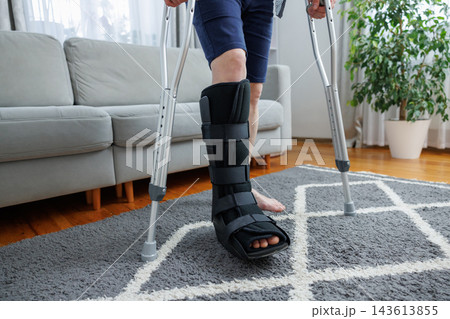 Person with leg injury using crutches and orthopedic boot at home Person with leg injury using crutches and orthopedic boot at home 143613855
