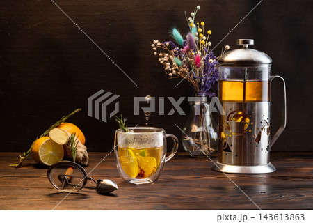 Dark mood tea still life with illuminated double wall glass cup, tea drops frozen in air, french press, dry flowers, ginger root, and spices on dark wooden table. Dark mood tea still life with illuminated double wall glass cup, tea drops frozen in air, french press, dry flowers, ginger root, and spices on dark wooden table. 143613863