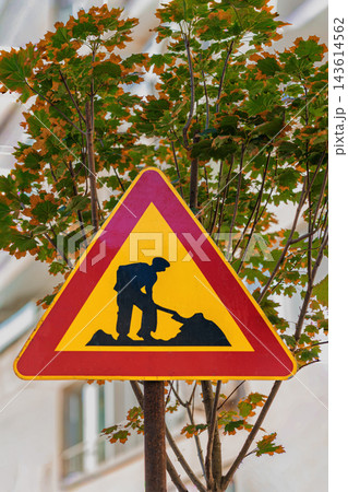 Triangular road work sign in front of autumn maple tree. Close-up of yellow and red road work warning traffic sign standing in front of colorful autumn tree leaves outdoors Triangular road work sign in front of autumn maple tree. Close-up of yellow and red road work warning traffic sign standing in front of colorful autumn tree leaves outdoors 143614562