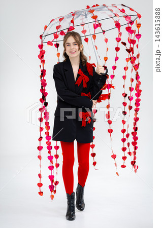 Young woman holding transparent umbrella decorated with hanging red heart garlands wearing black blazer with red bows and red tights Young woman holding transparent umbrella decorated with hanging red heart garlands wearing black blazer with red bows and red tights 143615888