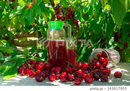 Jug of cherry juice and ripe cherries on table under tree leaves Jug of cherry juice and ripe cherries on table under tree leaves 143617953