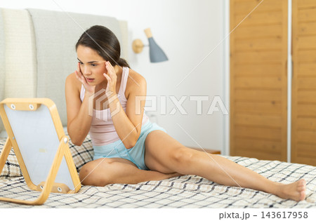 Attentive young girl looking at her face in mirror in bedroom Attentive young girl looking at her face in mirror in bedroom 143617958