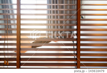 Wooden Venetian blinds separating a hotel bathroom and bedroom with a woman sleeping under a white blanket in the morning, providing privacy, light control and a peaceful atmosphere. 143618479