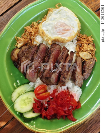 Appetizing Japanese sirloin steak donburi featuring sliced steak rice fried egg pickled vegetables and fried garlic on a vibrant green plate overhead view. Appetizing Japanese sirloin steak donburi featuring sliced steak rice fried egg pickled vegetables and fried garlic on a vibrant green plate overhead view. 143618561